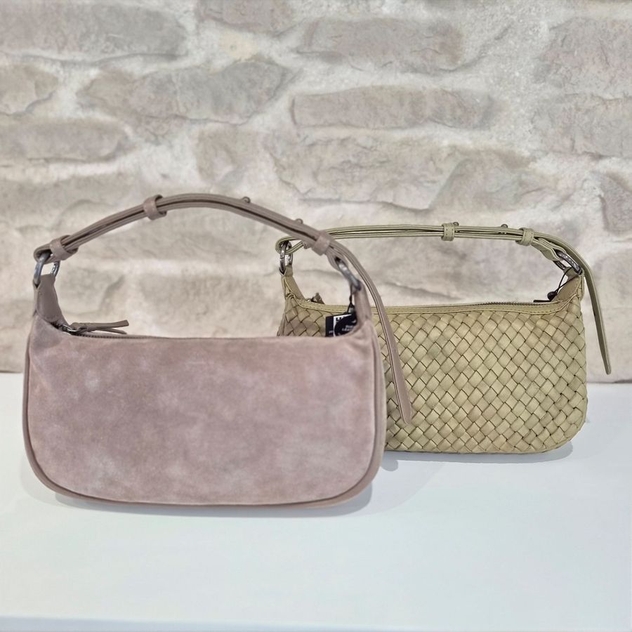 Zwei kleine Handtaschen mit kurzen Henkeln, eine aus glattem Veloursleder, die andere mit geflochtenem Muster, vor einer hellen Steinwand